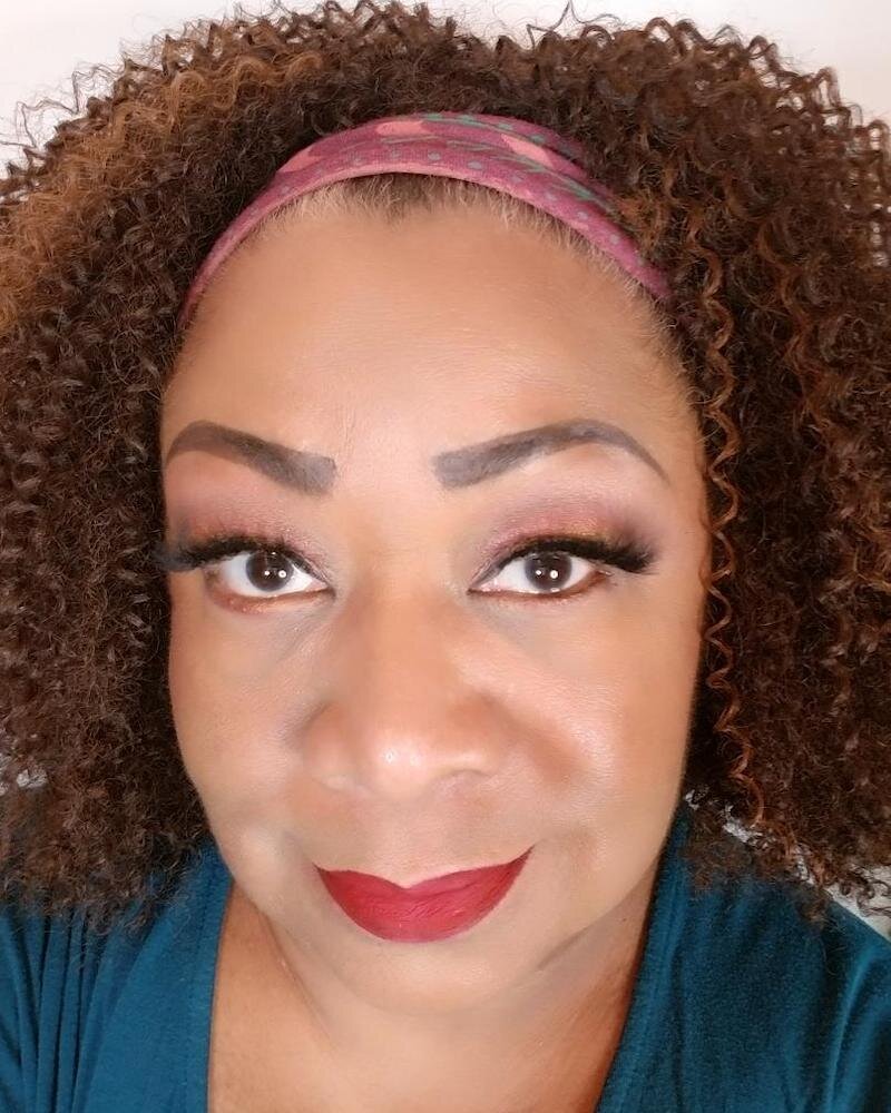 Pam's face fills the frame with tight brown spiral curls framed by a magenta print headband. While her makeup isn't excessive she's proud of her evenly applied lashes and bright red lip that enhances her impish smile,