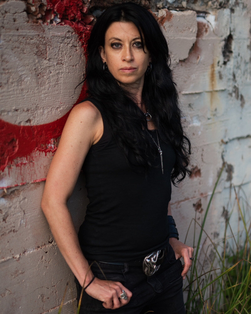 Photo of Carson Beker (they/them). A queer white person with long black hair and tattoos leans against a tumbledown wall. They are facing the camera and half smiling.