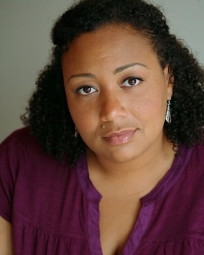 A headshot photo of Genevieve Jessee, a caramel skin toned Black woman with brown-black 3C curls, looks directly at the camera appearing to withhold a smile. Parts of her shoulders and neck&nbsp; outfitted in a dark purple shirt can be seen along wi…