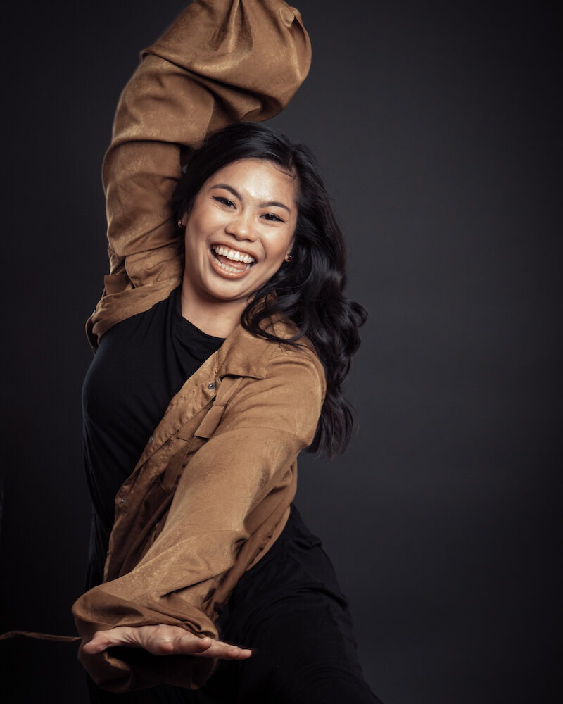 Action shot of Nikki Meñez, a Filipinx femme with honey-brown skin and curled, medium length, black hair. They are leaping joyfully to the viewer-left side of the frame wearing a tan jacket, a black shirt, black pants, and a wide grin.