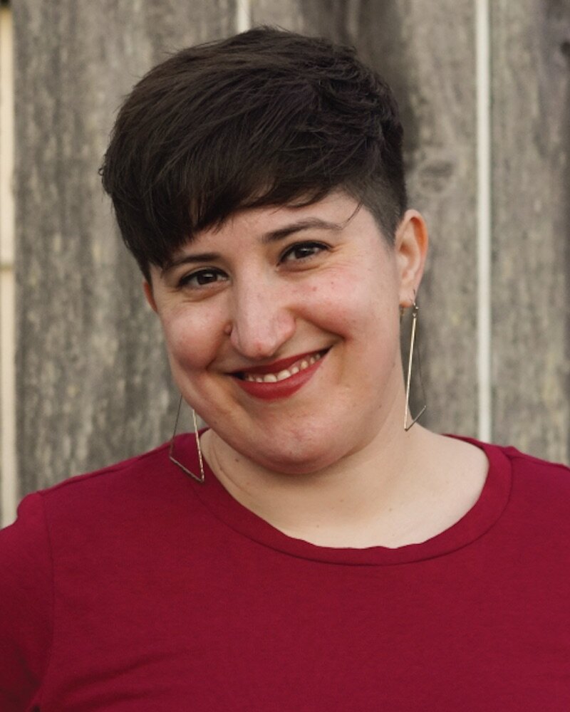 Photo of Nicole Jost (she/her), a white woman with short brown hair, smiling at the camera. She wears a dark red shirt, matching lipstick, and gold triangle earrings. Her expression is soft and inviting. Behind her, a weathered wooden fence.