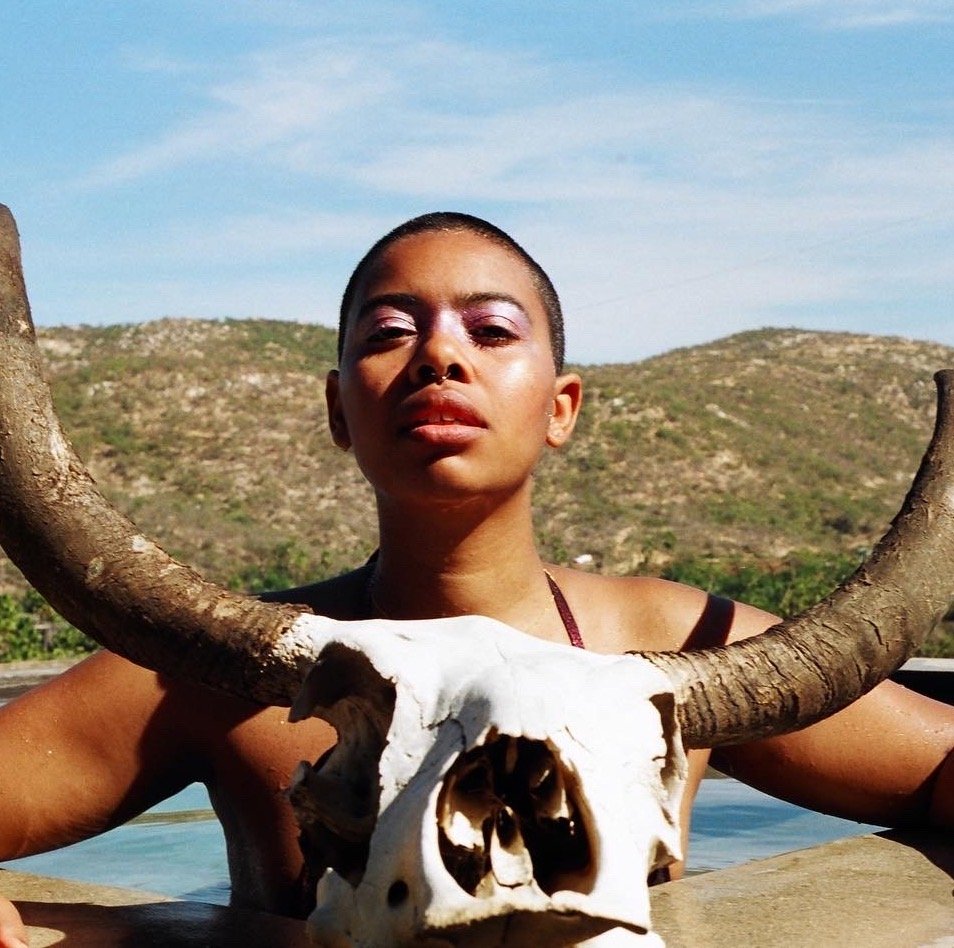 Portrait of Creatrx, a Black nonbinary artist, here shown emerging from a body of water with a large, horned, animal skull. They stare directly at the camera, unsmiling, magical.