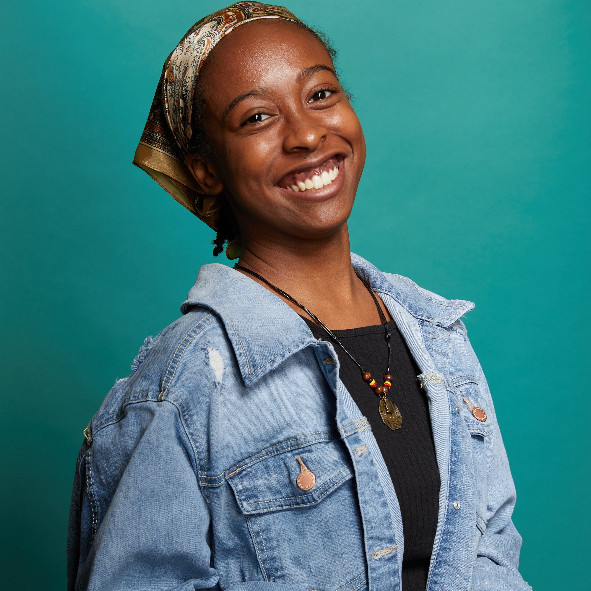 Headshot of storyteller/artist Tykaia Riley, a Black woman wearing a jean jacket and scarf over her head, she is tilting her head back and smiling wide, looking straight at the camera in a way that conveys confidence and joy.