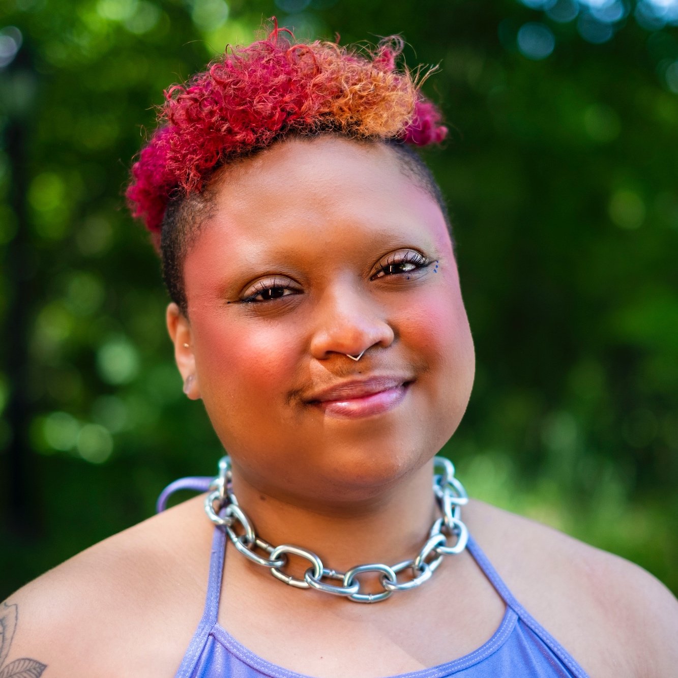 Headshot of the artist x (photo cred Friday Anderson), fae is a Black person with short curly magenta and orange hair, here shown wearing a shiny silver chain and a periwinkle top and rosy makeup, smiling at the camera head on.