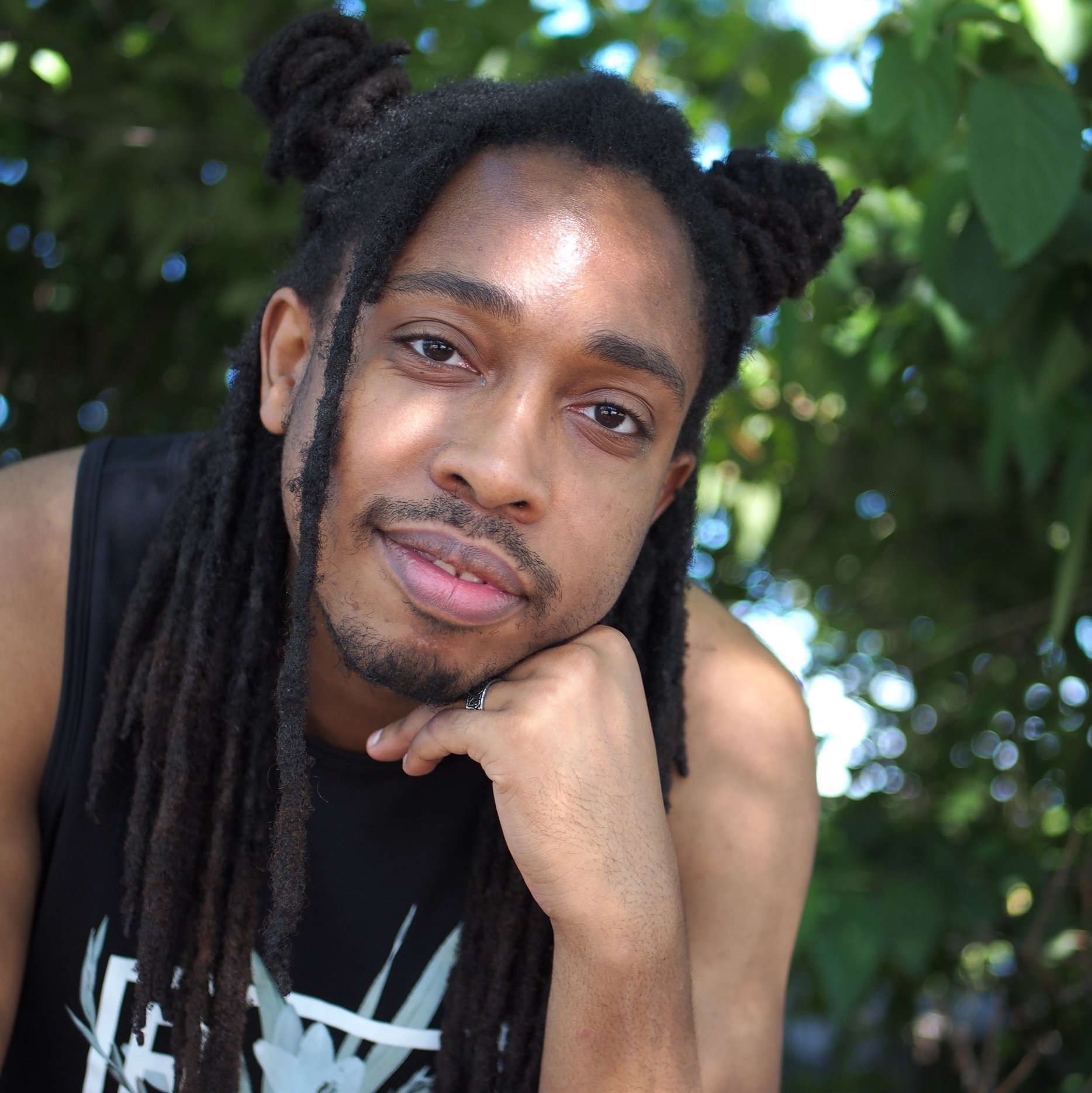 Headshot of the playwright Devin Randall, a Black man with long dreads shown outside under lush trees, he leans his head on one hand and leans forward with a half smile, his expression full of depth and curiosity.