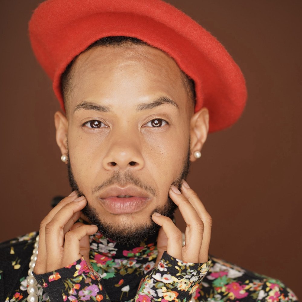 Headshot from shoulders up of Jaxin, a black, trans, queer, non-binary person with a beard. Jaxin is wearing a floral print turtle neck, a red beret, as well as pearls.  Jaxin gentlyJaxin gently holds their face with finger tips touching each cheek.