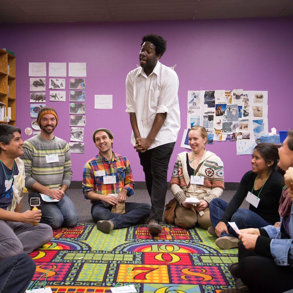 Portrait of the artist e. Franklin, a Black artist wearing a white shirt and black pants, here shown teaching a workshop in a bright room, they are smiling and moving, and everyone around them is smiling or laughing.