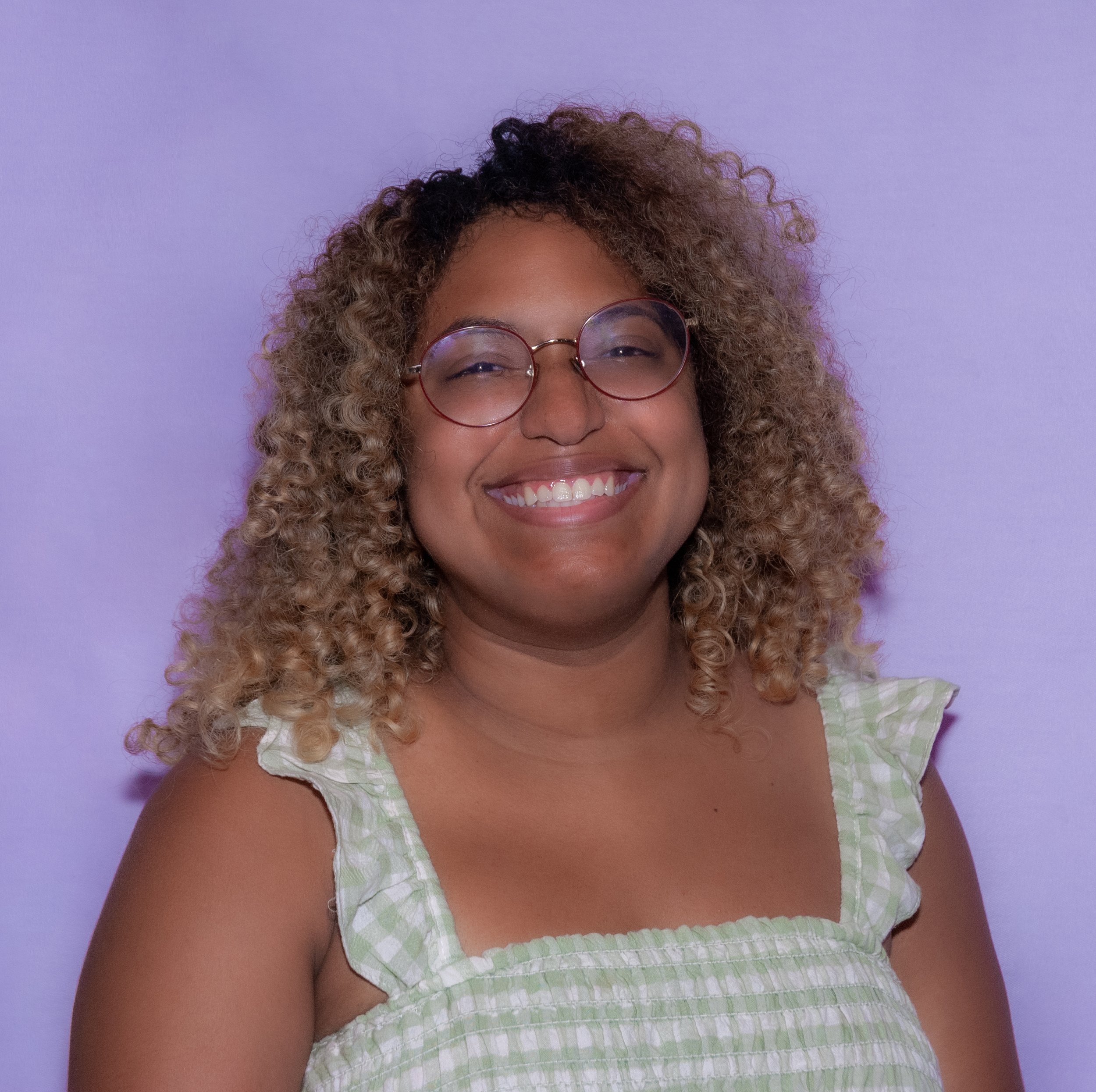 Portrait of playwright and director Kendall Philips, a Black queer person with curly light hair wearing round gold glasses, a checkered dress, and smiling wide directly at the camera, letting the smile take over their whole face.
