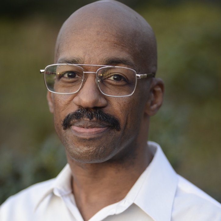 Portrait of the artist Louis Devaughn "DeVo" Nelson, a Black man here bald with a lush mustache, here wearing silver thin glasses and a white button up shirt, looking into the camera, almost smiling, his expression strong and kind.