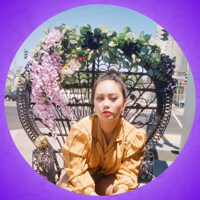 Portrait of Kayla May sitting outside in a Peacock chair adorned with plants. KMPS is wearing a yellow top and leaning forward, looking into the camera.