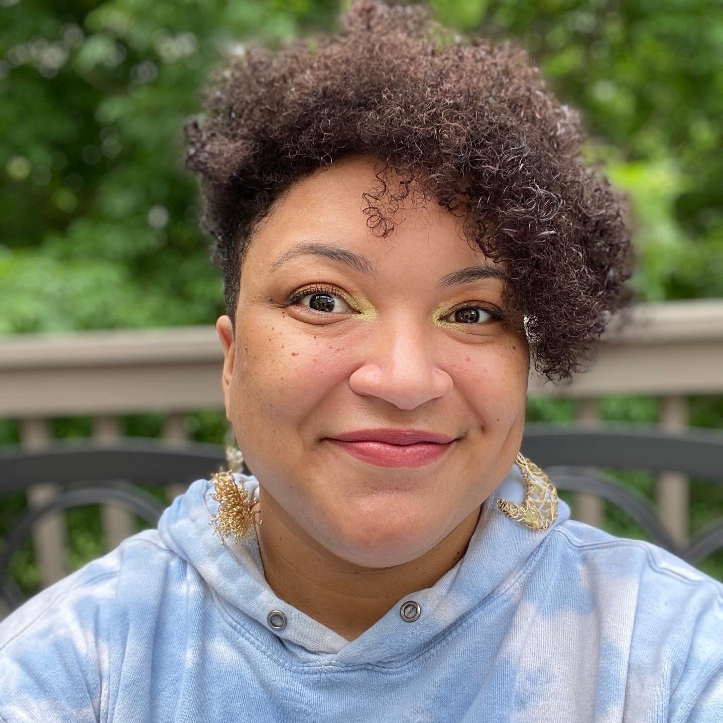 Headshot of the multi-disciplinary artist Artemis Montague, a Black mixed-race nonbinary creator, here shown wearing a sweater like a blue sky with clouds, smiling straight at the camera, with bright gold earrings, eyeshadow, and a dynamic smile.