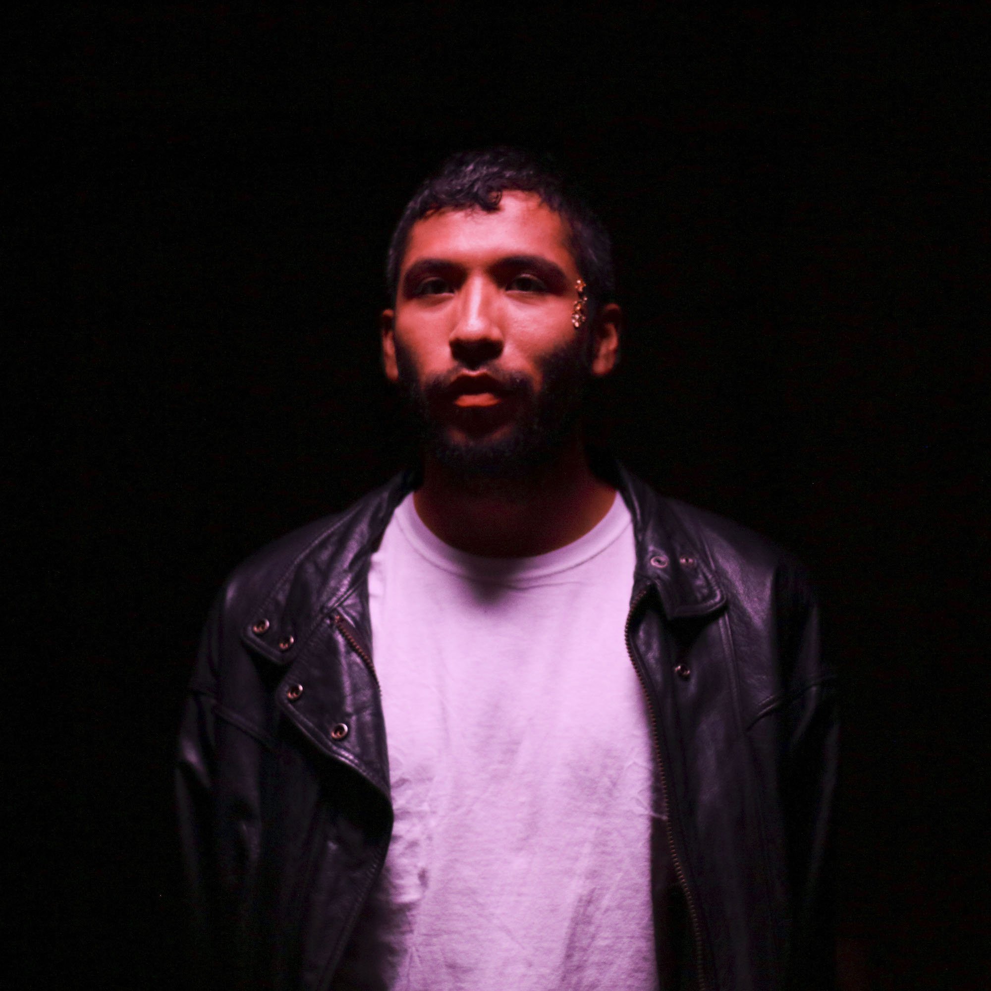 Headshot of the artist Estrellx Supernova, they are wearing a white shirt and black leather jacket, and stand in the dark illuminated by a warm horizon that lights up their face.