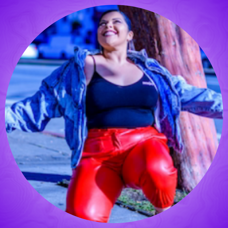 Portrait of Jessica kneeling outside on the sidewalk, her arms outstretched and looking up to the sky. She's wearing a black spaghetti strap tank top, red pants, and a denim jacket.