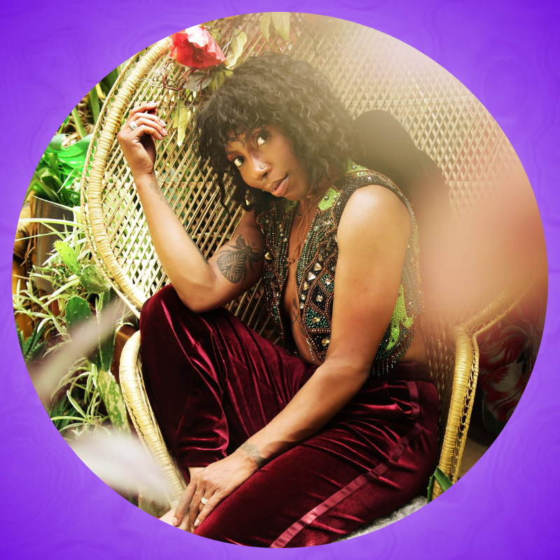 A caramel skinned Afro-Boricua with loose curly hair, tattoos, and gold facial piercings wears burgundy velvet pants and a green vest bedazzled with sparkling glass. They are sitting in a wicker Peacock chair surrounded by Moroccan rugs and plants.