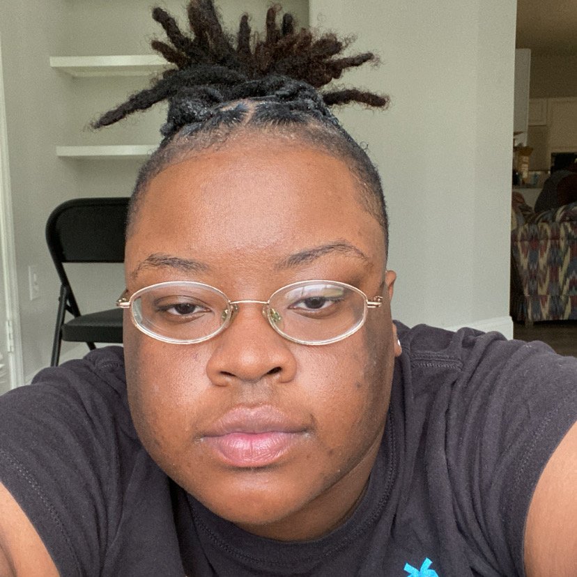 Portrait of the artist and opera singer Teddy Cozee Smith, a Black person wearing a black shirt and oval glasses, hair up, facing the camera, with a look that conveys intensity, peace, and focus.