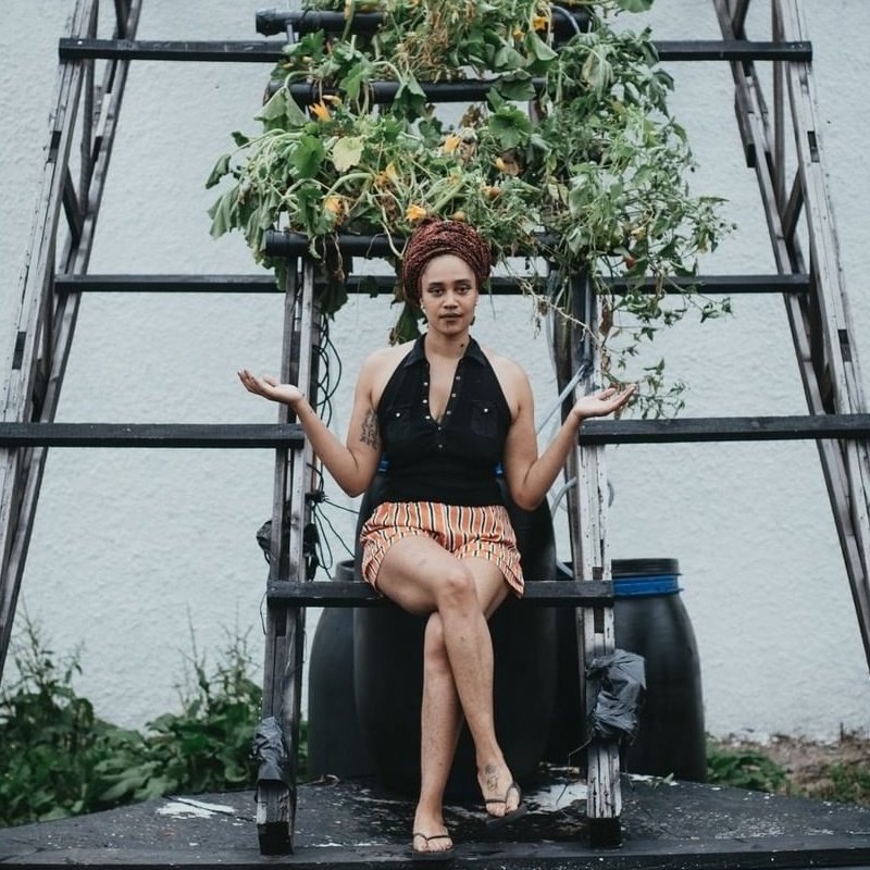 Portrait of the artist JNK Enzo, they are a Black person wearing bright striped shorts and a Black v necked top, and an Earth toned head wrap. They sit under a ladder, trellis dripping with flowers, palms out, smiling serenely.