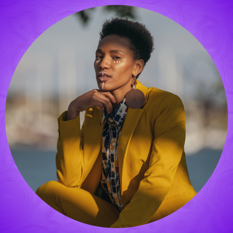 Janesta poses in a crouched position in a yellow tailored suit with a multicolored patterned button down shirt with white accent makeup looking straight forward.