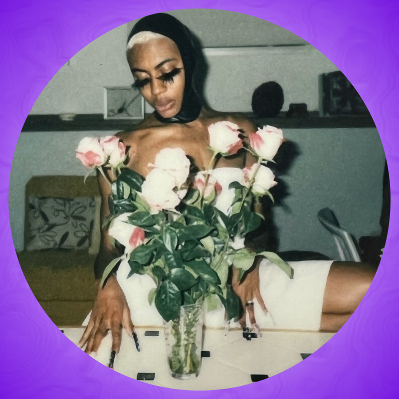 An image of Dillon wearing a black latex open faced hood, white dress strapless dress posing behind a vase filled with pink and white roses.