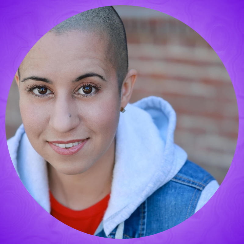 Actor, Edna Mira Raia (she/her), seated against a blurry brick background. With an open mouth smile, she looks slightly up at the lens, sporting a bright red t-shirt and jean jacket with gray cotton hood and sleeves.