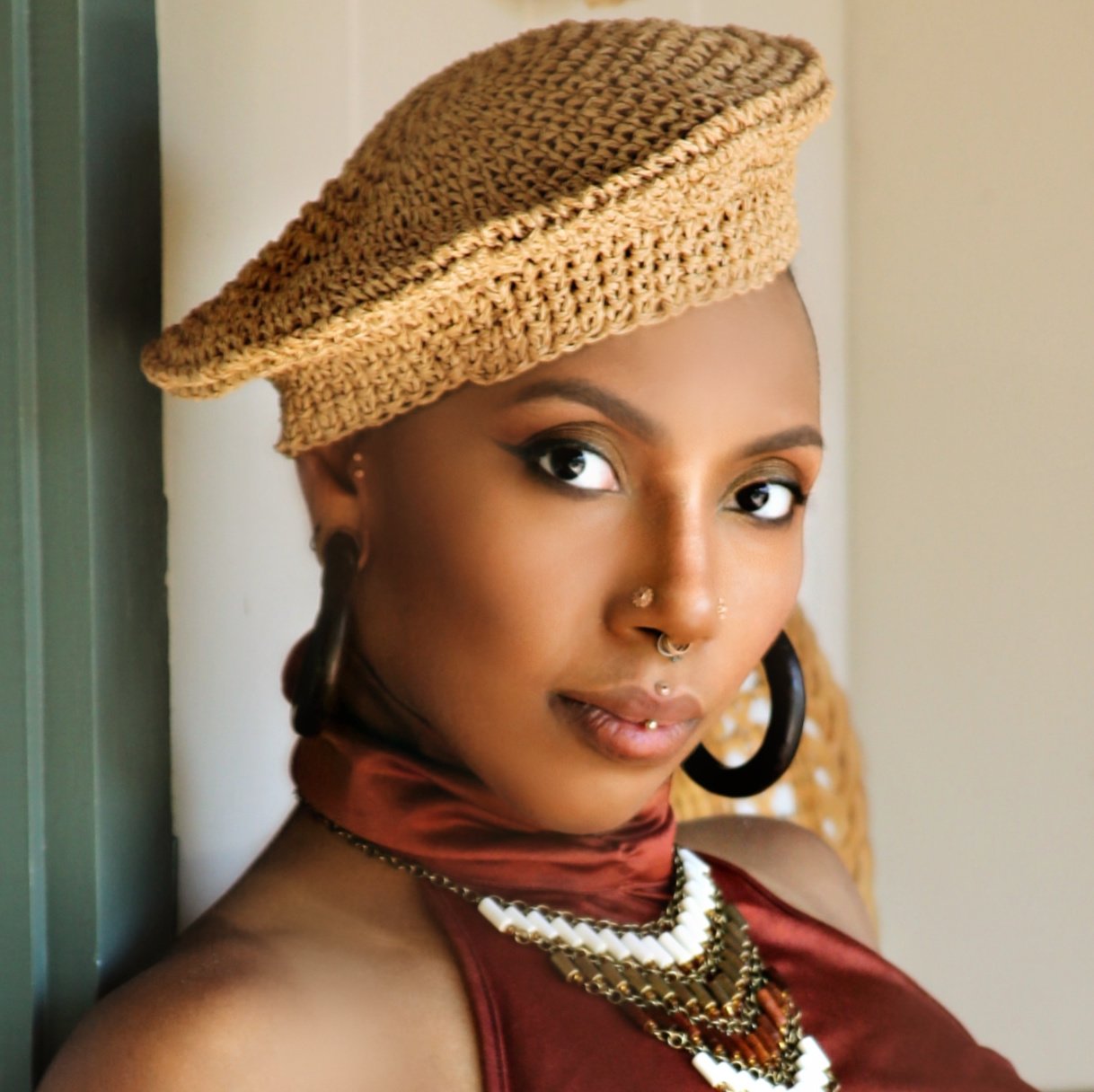 A headshot of Jes DeVille, a brown-skinned person with a knitted hat and a high-neck red shirt with multiple nose and ear piercings.