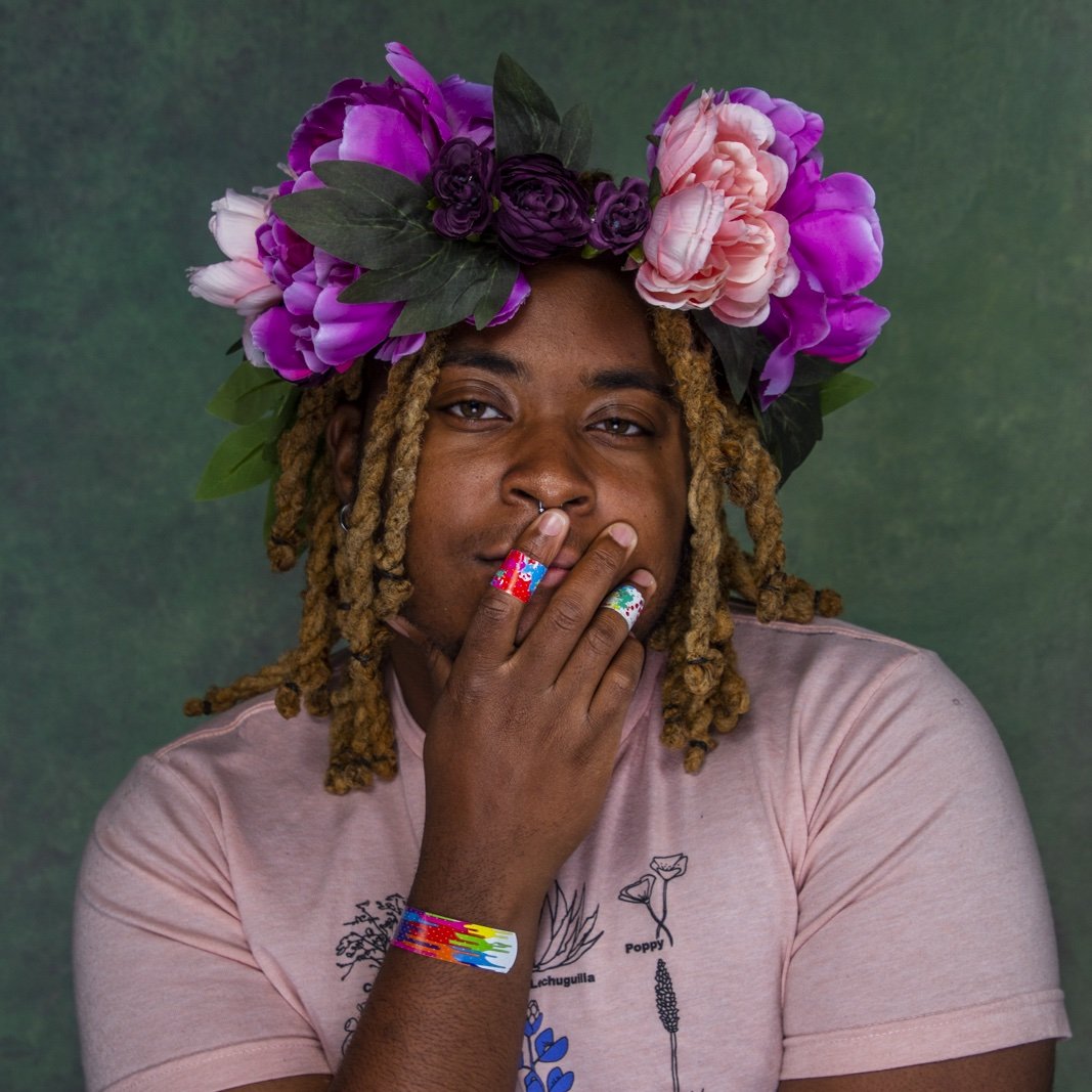 a Black person with brown skin — has a green background. They are wearing a flower crown, with pink and purple flowers, along with green leaves. Their hair is braided and blond. Their hand is slightly covering their mouth.